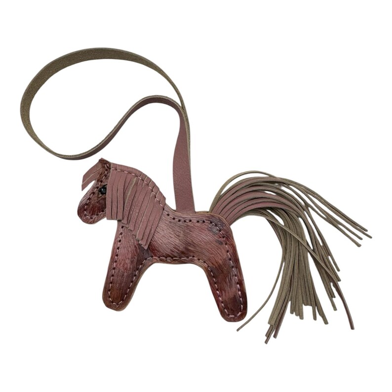 Op de afbeelding: Een leren paardenhanger in bruine en mauve tinten. Het paard heeft een gefranjerde manen en een lange, vloeiende staart. Er is een leren lus bevestigd om op te hangen. Deze hanger is waarschijnlijk een decoratief accessoire.