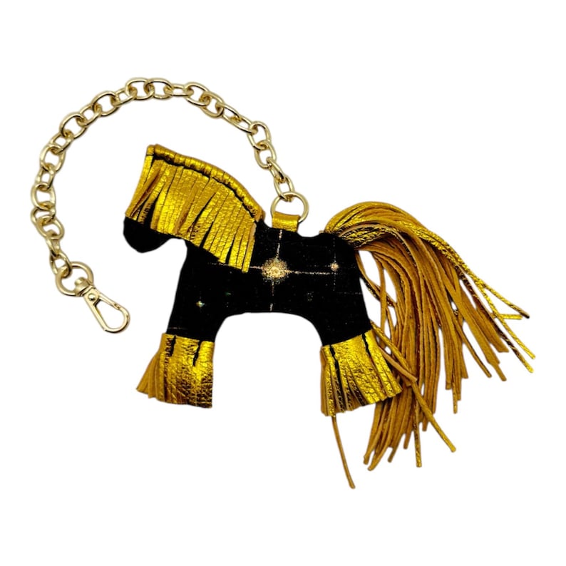 Op de afbeelding: Een zwart paardvormig sleutelhanger met gouden franje manen, staart en accenten. De sleutelhanger heeft een gouden ketting en sluiting. Het paard heeft een sterrenpatroon op zijn lichaam.