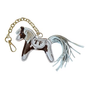 Op de afbeelding: Een leren paarden sleutelhanger met een bruin en wit patroon, een gouden ketting en een sluiting. Het paard heeft een witte manen en staart, en de initialen "TF" zijn op de zijkant gestikt. De sleutelhanger is ontworpen om aan sleutels of een tas te bevestigen.