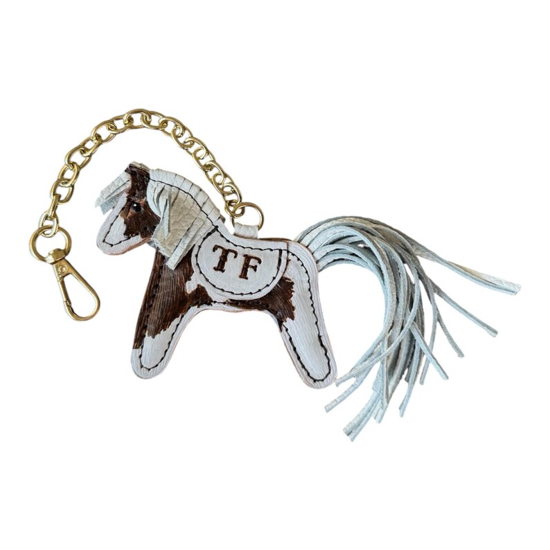 Op de afbeelding: Een leren paarden sleutelhanger met een bruin en wit patroon, een gouden ketting en een sluiting. Het paard heeft een witte manen en staart, en de initialen "TF" zijn op de zijkant gestikt. De sleutelhanger is ontworpen om aan sleutels of een tas te bevestigen.
