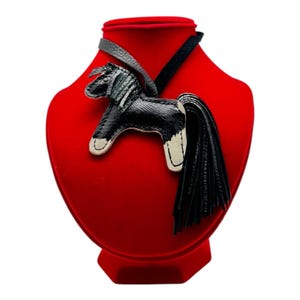 Op de afbeelding: Zwarte en witte leren paardenhanger met lange staart, tentoongesteld op een rode fluwelen buste. Het paard heeft een gedetailleerde manen en is bevestigd aan een zwart leren koord. Een uniek accessoire.