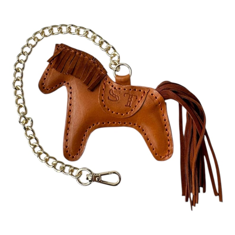 Puede incluir: Un llavero de cuero marr&oacute;n en forma de caballo con una cadena dorada y una cola de flecos. El caballo tiene las iniciales "ST" grabadas en su costado.