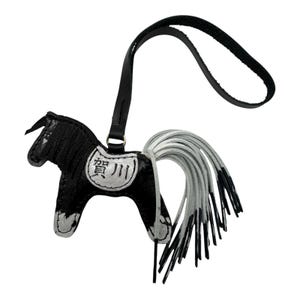 Op de afbeelding: Een zwart-witte leren paardenhanger met een lange, vloeiende witte staart. Het paard heeft een zwarte riem en een kleine metalen ring. De hanger heeft Japanse tekst.
