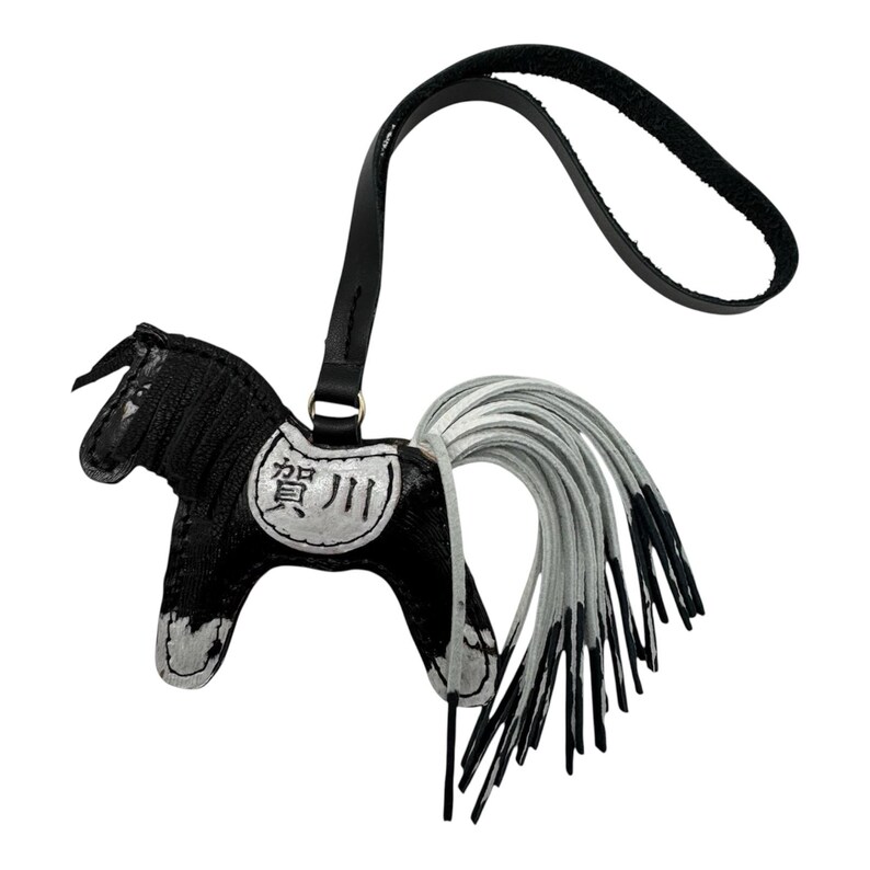 Op de afbeelding: Een zwart-witte leren paardenhanger met een lange, vloeiende witte staart. Het paard heeft een zwarte riem en een kleine metalen ring. De hanger heeft Japanse tekst.