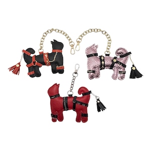 May include: Three animal-shaped keychains in black, metallic pink, and red. Each features a gold or silver chain, black or red straps with small metal hardware, and a matching tassel attached to the tail. The figures have a textured finish.