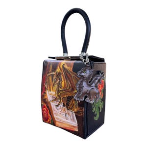 May include: Black handbag with a dragon and book design. The bag features a black handle, a dragon charm, and a detailed illustration of a dragon on a book with roses. The bag has a rectangular shape.