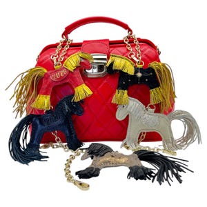 Op de afbeelding: Een rode gewatteerde handtas met een handvat aan de bovenkant en een gouden kettingriem. Vijf paardvormige sleutelhangers in verschillende kleuren, waaronder rood, zwart en zilver, hangen aan de tas. De rode paardensleutelhanger heeft het nummer "2026" erop.