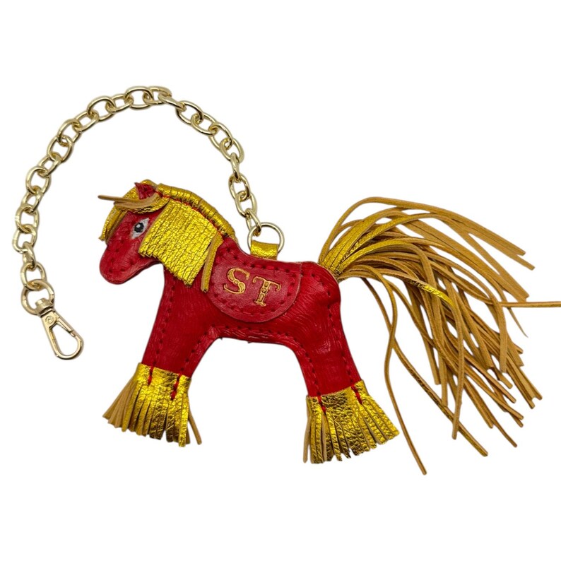 Op de afbeelding: Een rode leren paarden sleutelhanger met gouden accenten. Het paard heeft een gouden manen, staart en franjes op de benen. Het zadel heeft de letters "ST" erop geborduurd. Een gouden ketting en sluiting zijn bevestigd.