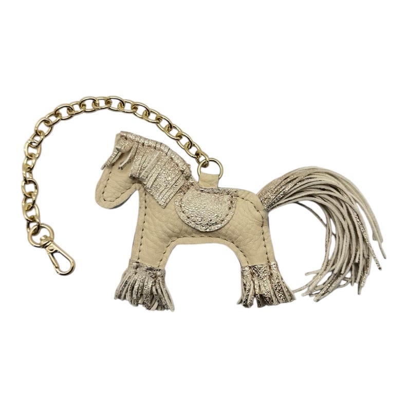 Op de afbeelding: Een beige leren paarden sleutelhanger met gouden accenten. Het paard heeft een gouden zadel, manen en franjes op de benen. Een gouden ketting en sluiting zijn bevestigd. Een accessoire met paardenthema voor sleutels of tassen.