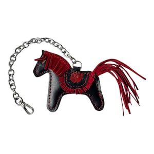Puede incluir: Un llavero de cuero negro en forma de caballo con una crin y una cola de flecos rojos. El llavero est&aacute; unido a una cadena plateada.