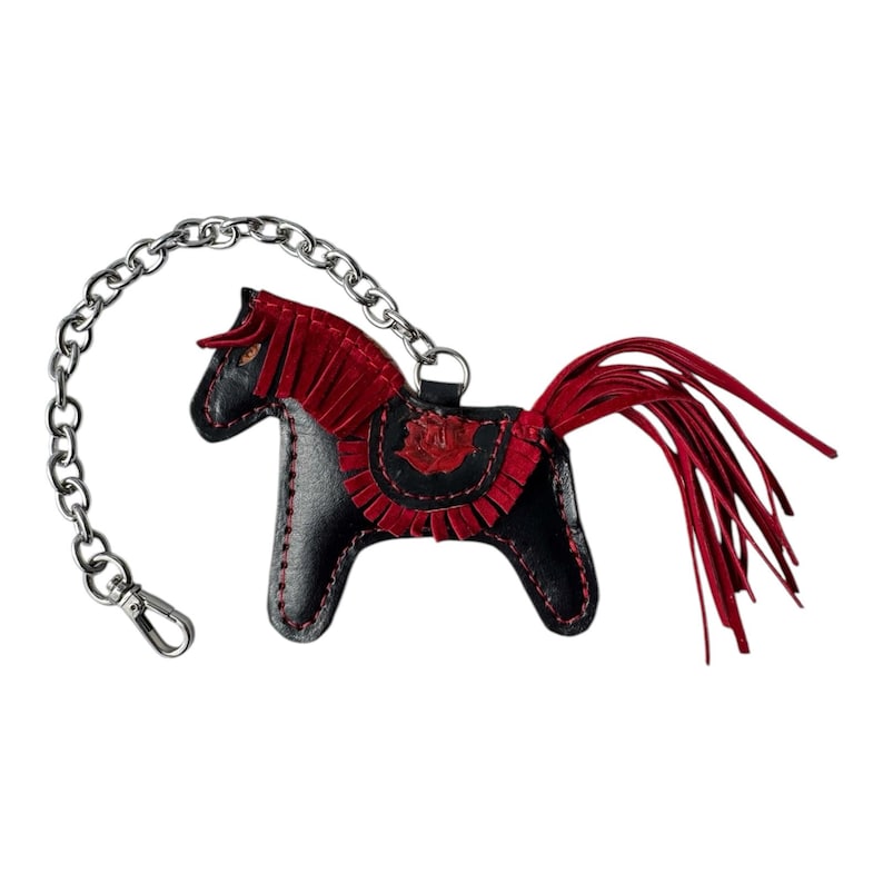 Puede incluir: Un llavero de cuero negro en forma de caballo con una crin y una cola de flecos rojos. El llavero est&aacute; unido a una cadena plateada.