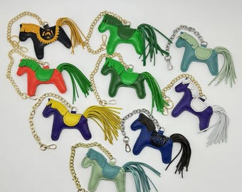 May include: Assortment of colorful leather horse keychains with gold chains and tassels. Each keychain features a different color horse with a saddle and a tassel tail.