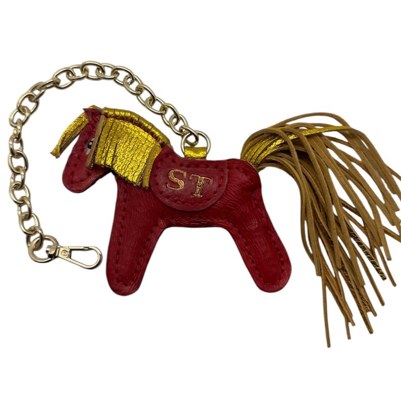 Op de afbeelding: Een rode leren paarden sleutelhanger met gouden accenten op de manen en staart. Het paard heeft een gouden zadel met de initialen "ST" en is bevestigd aan een zilveren ketting met een sluiting.