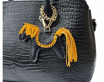 May include: Black leather handbag with a textured crocodile pattern. A gold chain and a horse-shaped leather charm with yellow fringe and accents hang from the handle. The charm is black with yellow details.