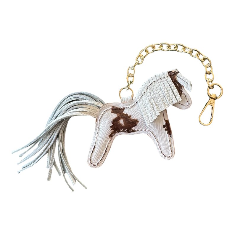 Op de afbeelding: Een witte en bruine leren paarden sleutelhanger met een gouden ketting en sluiting. Het paard heeft een witte manen en staart, en bruine vlekken op zijn lichaam. Een decoratief accessoire.