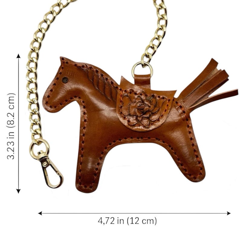 May include: A brown leather horse keyring with a gold chain. The horse has a saddle with a floral design and a fringed tail. The keyring is 4.72 inches (12 cm) wide and 3.23 inches (8.2 cm) tall.