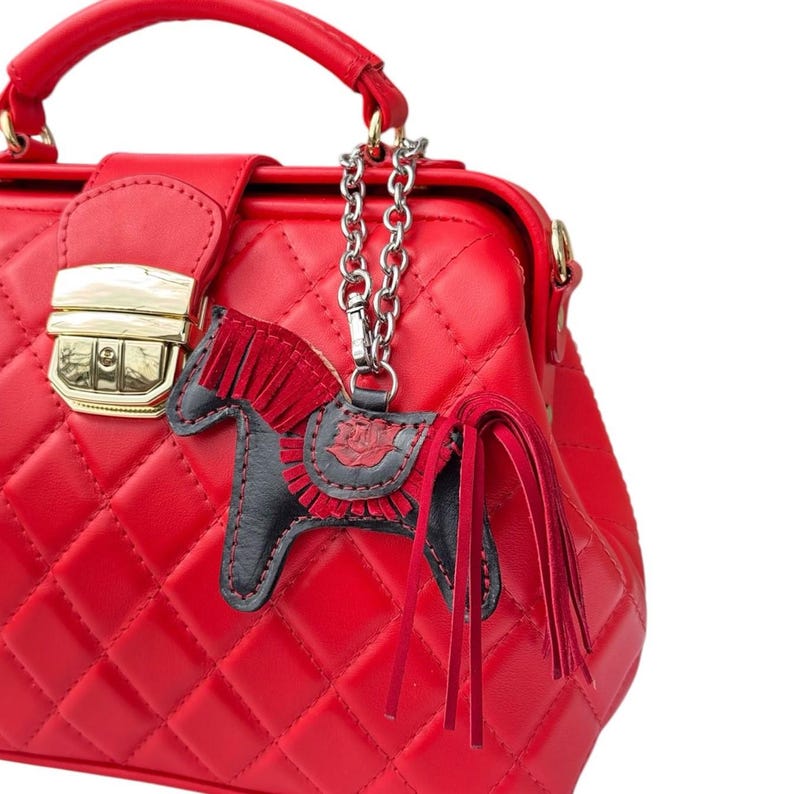 Puede incluir: Un bolso de mano de cuero acolchado rojo con cierre dorado y un dije de caballo de cuero negro colgando de una cadena plateada.