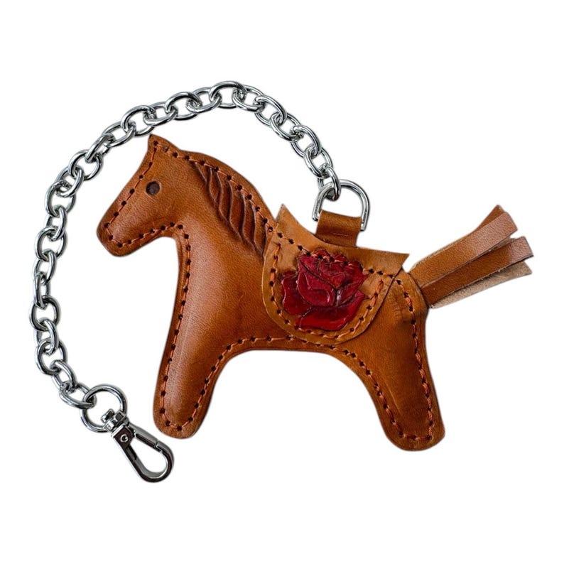 May include: A brown leather horse keyring with a red rose design on the saddle. The keyring is attached to a silver chain.