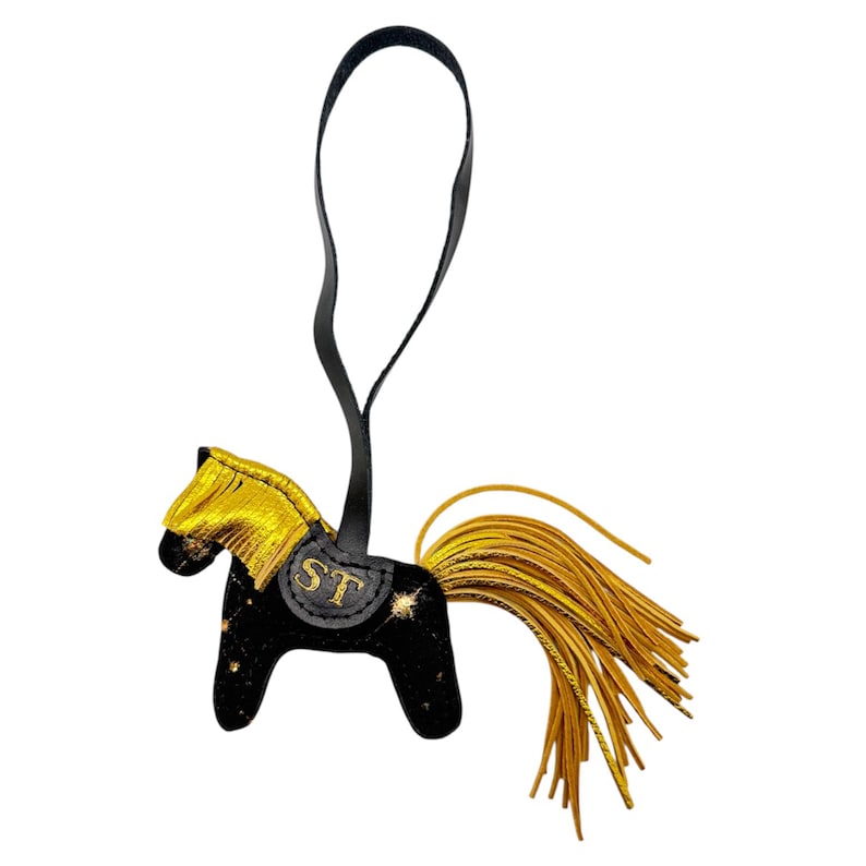 Op de afbeelding: Een zwart en goudkleurig paardvormig bedeltje met een zwarte leren riem. Het paard heeft een gouden manen en staart, met de letters "ST" op het zadel. De bedel is versierd met gouden sterachtige accenten.