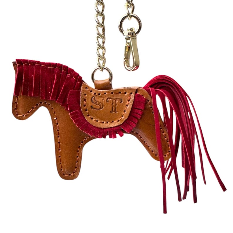 Puede incluir: Un llavero de cuero marr&oacute;n en forma de caballo con una crin y una cola de flecos rojos. El llavero tiene una cadena dorada y un cierre. El caballo tiene las iniciales "ST" grabadas en su silla de montar.