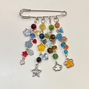 May include: A decorative safety pin with dangling charms. The charms include colorful beads, stars, hearts, and cloud shapes. The pin is silver-toned, and the charms are in various colors, including red, blue, yellow, and green.