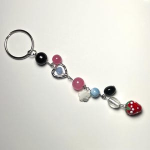 May include: A beaded keyring with a silver ring. The keyring features pink, black, white, and blue beads, a silver heart charm, and a red strawberry charm with white dots.