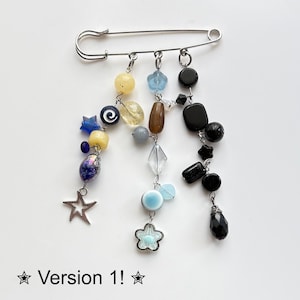 May include: A large silver safety pin with dangling beaded charms. The charms include yellow, blue, brown, and black beads, along with a silver star and a flower. The text "Version 1!" is at the bottom.