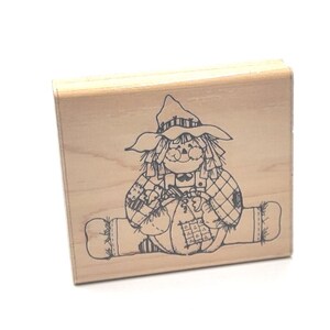 May include: A rectangular wooden rubber stamp featuring a black line art illustration of a scarecrow wearing a wide-brimmed hat and patched clothing, sitting while holding a large pumpkin. The stamp has a light-colored natural wood block base.