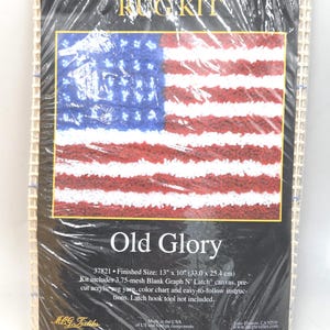 May include: A Graph N' Latch rug kit featuring an American flag design, titled "Old Glory." The finished size is 13" x 10". The kit includes a canvas, pre-cut acrylic rug yarn, and instructions. The flag design has red and white stripes with a blue field of stars.