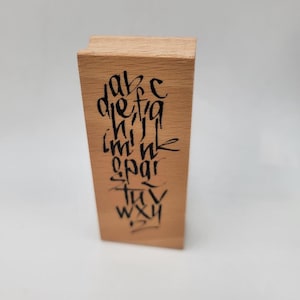 May include: A rectangular wooden stamp with black calligraphy lettering. The letters are arranged vertically and include the alphabet. The wood has a natural grain and a light brown color. The stamp is set against a white background.
