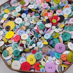 May include: A large collection of assorted sewing buttons in a variety of colors, shapes, and sizes, including round, flat, and multi-hole designs, contained within a hexagonal metal container with a copper-toned rim.