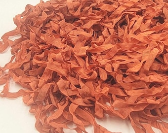 May include: A large pile of thin, rust-colored fabric strips with a slightly wrinkled and textured appearance. The strips are loosely tangled together, creating a dense, layered mass of narrow, flexible material with frayed edges throughout.