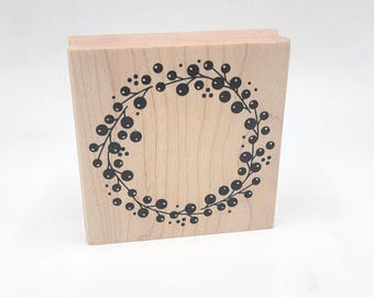 May include: A wooden stamp featuring a wreath design with black berry-like details. The stamp is made of light-colored wood and is rectangular in shape. The wreath is circular and surrounds a blank space.