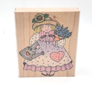 May include: A rectangular wooden rubber stamp featuring a line art illustration of a person wearing a straw hat with a sunflower, holding a potted plant and a watering can. The person wears a dress with a pink heart patch and a large bow.