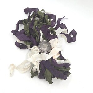 May include: A handmade fabric flower with a metal star button center. The flower features layers of ruffled fabric in shades of purple, olive green, and cream. This unique accessory can be used as a hair clip or brooch.