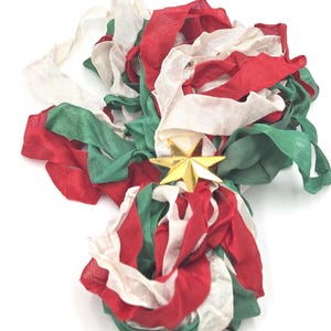 May include: A bundle of sheer fabric ribbons in red, white, and green, gathered together and secured in the center by a shiny, metallic gold-colored six-pointed star ornament. The ribbons have a loose, flowing texture and are layered together.