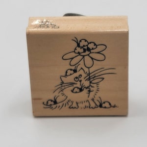 May include: A wooden stamp featuring a cartoon cat holding a flower. The cat is drawn in black ink, with a whimsical expression. The stamp has a rectangular shape and a handle on top.