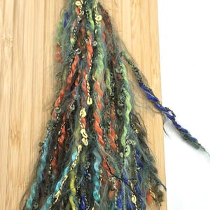 May include: A decorative tassel made of multi-colored yarn strands in shades of green, orange, blue, and yellow, featuring metallic gold accents and fuzzy textures. The tassel is attached to a bronze-colored metal lobster clasp at the top.