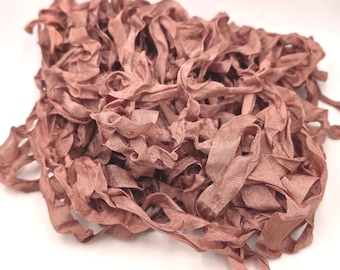 May include: A tangled pile of dusty rose colored ribbon with a visible woven texture. The ribbon is thin and flexible, featuring frayed edges and a matte finish, bunched together in a loose, irregular heap against a plain white background.