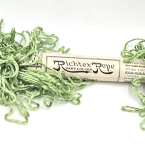May include: A bundle of light green thread wrapped in a paper label that reads Richtex Rope Fast Colors. The thread is loosely coiled on both sides of the central paper band, which also contains partial printed text about artificial silk.
