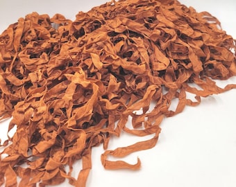 May include: A pile of rust-colored fabric strips, possibly ribbon or trim, with a slightly ruffled texture. The strips are scattered, creating a dense, textured appearance. The color is a warm, earthy tone.