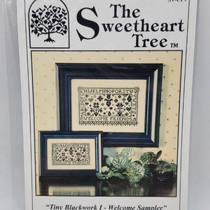 May include: A cross-stitch pattern for a sampler titled "Tiny Blackwork I - Welcome Sampler". The pattern features a black and white alphabet, a floral design, and the text "Welcome Friends". The pattern is designed by Sandra Cox Vanosd and is available from The Sweetheart Tree.