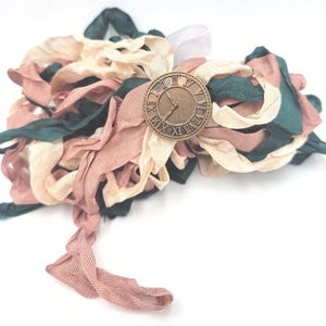 May include: A handmade fabric flower with a bronze clock face in the center. The flower is made of layered ribbons in shades of pink, cream, and green. The clock face has Roman numerals and hands pointing to the time.