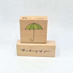 May include: Two wooden stamps. The top stamp features a green umbrella graphic. The bottom stamp has the phrase "thinking of you" in a cursive font. Both stamps are rectangular and made of light-colored wood.