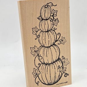May include: A rectangular wooden rubber stamp featuring a black ink line drawing of five stacked pumpkins. The design includes curling vines and leaves surrounding the pumpkins, with the text JEAN ARMENT 2000 printed near the bottom right corner.