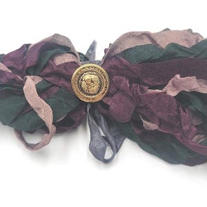 May include: A handmade hair accessory featuring layered fabric ribbons in shades of green, purple, and mauve. A gold-colored button with an embossed design secures the ribbons. The accessory is designed for hair styling.