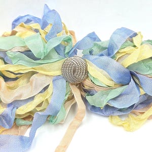 May include: A decorative cluster of thin, frayed fabric ribbons in pastel shades of light blue, pale yellow, soft green, and peach. The ribbons are gathered and secured in the center by a round, metallic button featuring a textured, radial pattern.