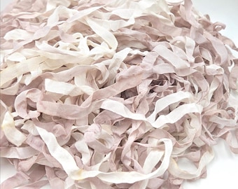 May include: A large pile of thin, narrow fabric ribbons in shades of dusty mauve and off-white. The ribbons have a soft, slightly wrinkled texture and are tangled together in a loose, voluminous heap, displaying varied lengths and widths.