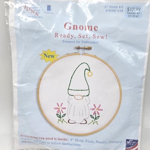 May include: A 6-inch embroidery hoop kit featuring a gnome design. The kit includes a stamped pattern, floss, and needle. The gnome is depicted with a green hat and a white beard, surrounded by pink flowers. The text "Gnome Ready, Set, Sew!" is displayed.