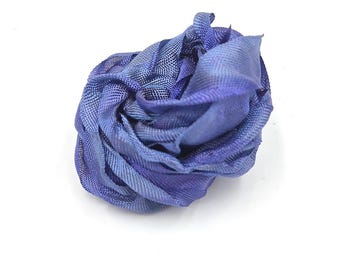 May include: A decorative flower accessory crafted from a single piece of blue mesh fabric. The material is folded and layered to create a dense, rounded shape with visible woven texture and varying shades of blue throughout the petals.